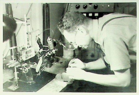 A black and white photograph of Sir Bernard Katz in their University College London lab. They are hunched over working at a bench near a microscope. Photograph from the UCL Special Collections.